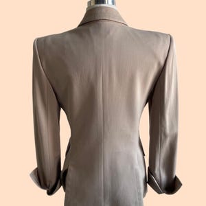 Pu&ograve; includere: Un blazer beige esposto su un manichino. Il blazer ha un design classico con revers a lancia, chiusura a due bottoni e maniche lunghe con polsini risvoltati. La vista posteriore mostra una vestibilit&agrave; sartoriale e una sottile trama nel tessuto.