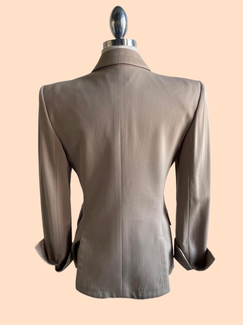 Pu&ograve; includere: Un blazer beige esposto su un manichino. Il blazer ha un design classico con revers a lancia, chiusura a due bottoni e maniche lunghe con polsini risvoltati. La vista posteriore mostra una vestibilit&agrave; sartoriale e una sottile trama nel tessuto.