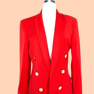 May include: A bright red double-breasted blazer with six cream-colored buttons. The blazer has a notched lapel and two front pockets. The garment is displayed on a white mannequin against a peach background.