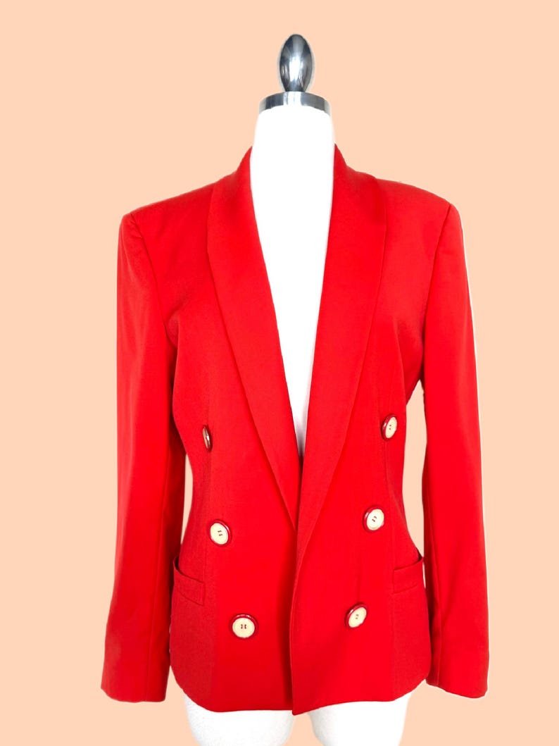 May include: A bright red double-breasted blazer with six cream-colored buttons. The blazer has a notched lapel and two front pockets. The garment is displayed on a white mannequin against a peach background.