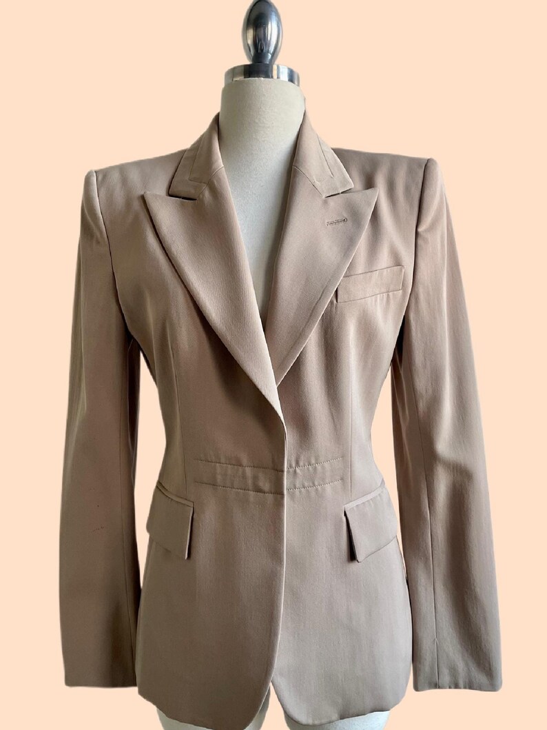 Pu&ograve; includere: Un blazer beige con revers a lancia, due tasche anteriori e chiusura a un bottone. La giacca su misura &egrave; esposta su un manichino su sfondo color pesca. Il blazer ha un design classico e professionale.
