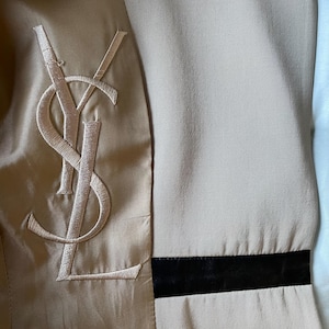 Pu&ograve; includere: Primo piano dell'interno di una giacca beige con un grande logo ricamato. La giacca presenta una fodera in raso e una fascia nera a contrasto. Il logo &egrave; un monogramma stilizzato in un colore beige pi&ugrave; chiaro.