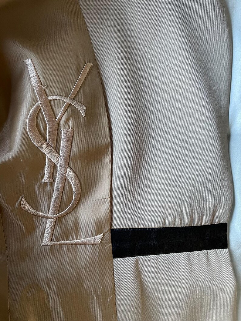 Pu&ograve; includere: Primo piano dell'interno di una giacca beige con un grande logo ricamato. La giacca presenta una fodera in raso e una fascia nera a contrasto. Il logo &egrave; un monogramma stilizzato in un colore beige pi&ugrave; chiaro.