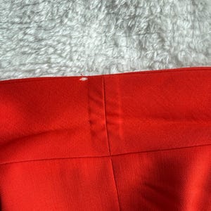 May include: Close-up of a section of bright red fabric, possibly clothing. The fabric has a smooth texture and is divided into panels by seams. A small white mark is visible near the top edge. The background is a fluffy, white surface.