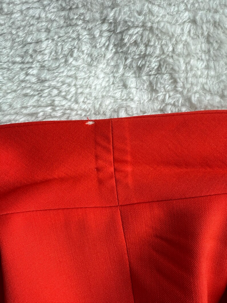 May include: Close-up of a section of bright red fabric, possibly clothing. The fabric has a smooth texture and is divided into panels by seams. A small white mark is visible near the top edge. The background is a fluffy, white surface.