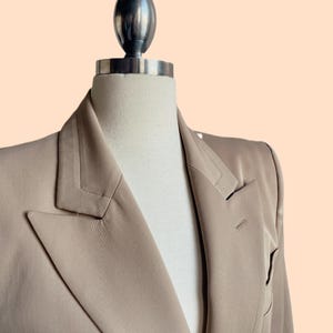 Pu&ograve; includere: Un blazer beige esposto su un manichino. Il blazer ha un revers a lancia, una chiusura a un bottone e maniche lunghe. Il tessuto &egrave; liscio e ha una leggera lucentezza. Il manichino ha una parte superiore argentata.