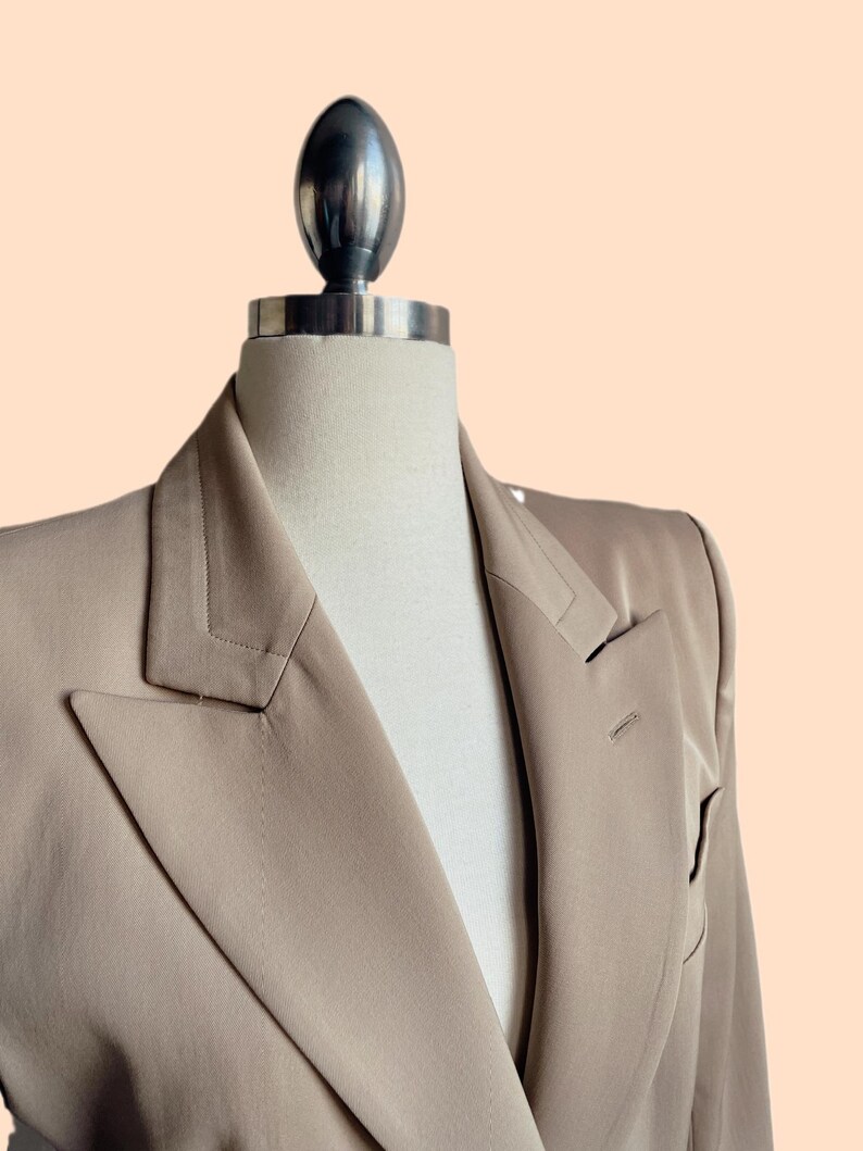 Pu&ograve; includere: Un blazer beige esposto su un manichino. Il blazer ha un revers a lancia, una chiusura a un bottone e maniche lunghe. Il tessuto &egrave; liscio e ha una leggera lucentezza. Il manichino ha una parte superiore argentata.