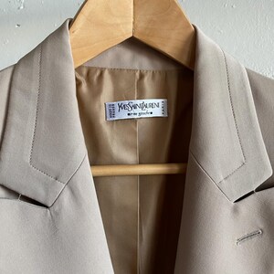 Pu&ograve; includere: Un blazer beige con revers a lancia e chiusura a un bottone. La giacca &egrave; appesa a un'appendiabiti di legno. L'etichetta all'interno della giacca recita "Yves Saint Laurent rive gauche".