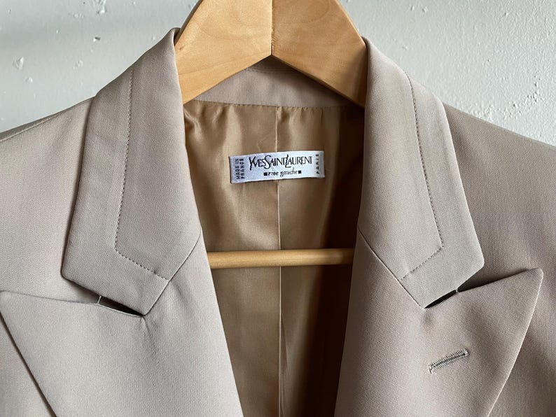 Pu&ograve; includere: Un blazer beige con revers a lancia e chiusura a un bottone. La giacca &egrave; appesa a un'appendiabiti di legno. L'etichetta all'interno della giacca recita "Yves Saint Laurent rive gauche".