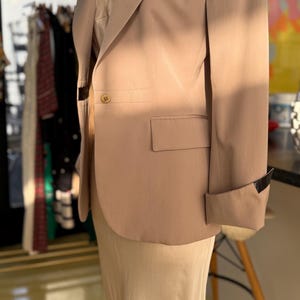 Pu&ograve; includere: Un blazer color beige con chiusura a un bottone e revers a lancia. Il blazer ha una tasca sul petto e un polsino ripiegato con un accento nero. &Egrave; indossato sopra un top e una gonna color crema.