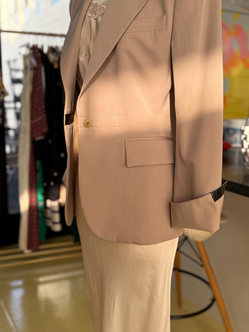 Pu&ograve; includere: Un blazer color beige con chiusura a un bottone e revers a lancia. Il blazer ha una tasca sul petto e un polsino ripiegato con un accento nero. &Egrave; indossato sopra un top e una gonna color crema.