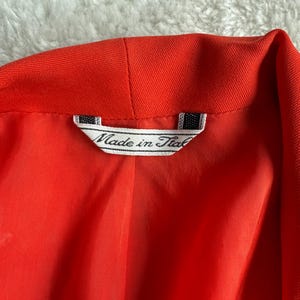 May include: Close-up of a bright red blazer's interior, showcasing the lining and collar. A tag reads "Made in Italy" in a classic font, indicating the garment's origin. The fabric appears smooth and the color is vibrant.