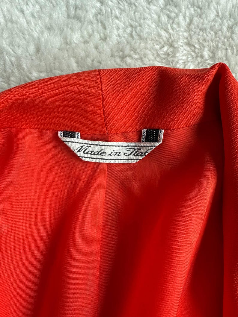May include: Close-up of a bright red blazer's interior, showcasing the lining and collar. A tag reads "Made in Italy" in a classic font, indicating the garment's origin. The fabric appears smooth and the color is vibrant.