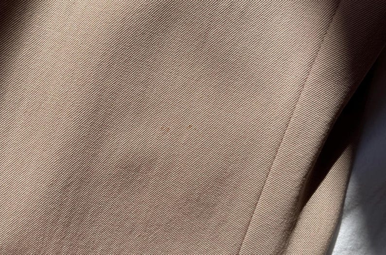 Pu&ograve; includere: Primo piano di una sezione di tessuto beige con una trama a coste diagonali. Il tessuto ha una leggera lucentezza e alcune piccole macchie pi&ugrave; scure. Il bordo del tessuto &egrave; visibile sul lato destro, con una cucitura.