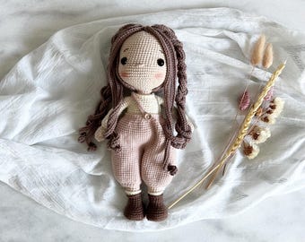May include: A crocheted doll with long brown hair, wearing a pink jumpsuit and brown boots. The doll is lying on a white fabric background.