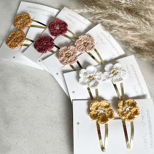 Könnte beinhalten: Satz mit fünf Paaren gehäkelter Blumenhaarspangen. Die Spangen sind in verschiedenen Farben, darunter Weiß, Gelb, Rosa und Rot. Jede Spange besteht aus zwei gehäkelten Blumen, die an einer goldenen Metallspange befestigt sind.