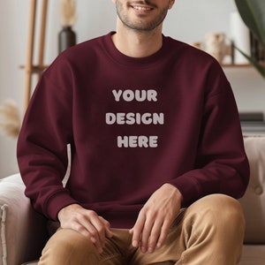 May include: A maroon sweatshirt with the text "YOUR DESIGN HERE" in white, block letters. The sweatshirt is a crew neck style and appears to be made of a soft material. The person is wearing khaki pants.
