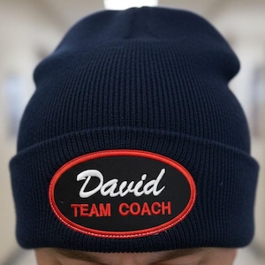 May include: Navy blue knit beanie with a black and red patch. The patch features the name "David" in white script above the words "TEAM COACH" in red. The beanie is a classic, casual style.