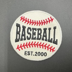 May include: A round, white baseball patch with red stitching and the word "BASEBALL" in black block letters. The text "EST. 2000" is below the word baseball. The patch has a white border.