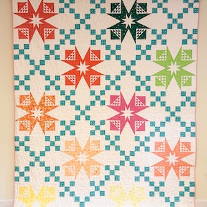 May include: A vibrant quilt with a star design. The quilt showcases large, multi-coloured stars in shades of red, green, orange, pink, and yellow, set against a white backdrop. The stars are linked by teal squares. The quilt is suspended from a wooden rod.