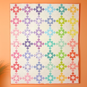 May include: A large, colourful quilt with a repeating pattern of star-shaped blocks in a rainbow of colours, including red, pink, purple, blue, green, and yellow. The quilt is displayed against an orange wall.