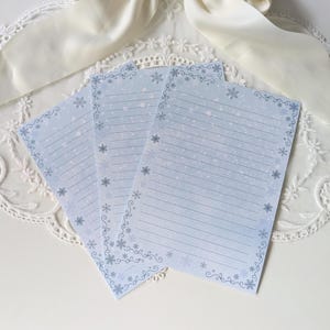 Könnte beinhalten: Drei hellblaue, linierte Briefpapierbögen mit einem Schneeflocken-Design. Das Papier hat einen Farbverlauf, der von Weiß zu Blau übergeht. Jedes Blatt hat einen dekorativen Rand mit Schneeflocken und Wirbeln. Das Briefpapier eignet sich zum Schreiben von Briefen oder Notizen.