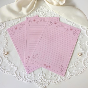 May include: Three pink lined notepads with floral and bow illustrations. The notepads have a decorative border and are arranged on a white lace surface. The notepads are a soft pink color.