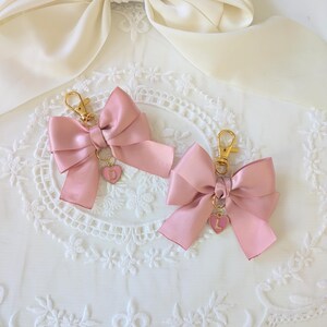 Peut inclure: Deux porte-cl&eacute;s en forme de n&oelig;ud rose satin&eacute; avec des attaches dor&eacute;es. Chaque n&oelig;ud est orn&eacute; d'un petit c&oelig;ur rose avec une lettre. Les porte-cl&eacute;s sont pr&eacute;sent&eacute;s sur une surface en dentelle blanche avec un ruban de satin cr&egrave;me en arri&egrave;re-plan.