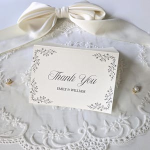 May include: A cream-coloured folded thank you card with black text reading Thank You and EMILY & WILLIAM, featuring delicate black floral vine corner illustrations. The card is placed on white lace fabric with a large cream satin bow above it.