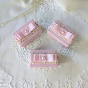 以下が含まれることがあります： 淡いピンク色の長方形のギフトボックス3つ。薄いピンク色のサテンリボン、パールアクセント、クリーム色のレーストリムが施されています。白いレースの表面に配置され、繊細でフェミニンな美しさを醸し出しています。小さなギフトに最適です。