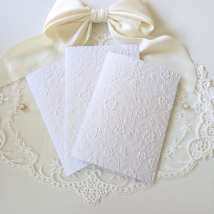 Puede incluir: Tres tarjetas blancas con relieve floral, dispuestas sobre una superficie de encaje blanco, con un gran lazo de satén color crema. Las tarjetas presentan un delicado patrón floral en relieve. Se ve un adorno de perlas y pedrería.