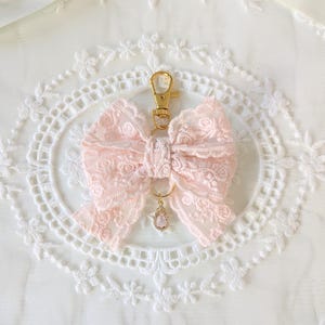 May include: A pink lace bow keyring with a gold-coloured clasp and a small, teardrop-shaped charm. The bow is intricately detailed, and the charm has a pink gemstone. The background is a white, lacy fabric.