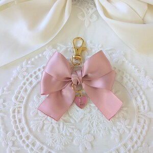 Peut inclure: Un porte-cl&eacute;s en forme de n&oelig;ud rose en satin, avec un fermoir dor&eacute; et un charm en forme de c&oelig;ur. Le charm porte la lettre "L". Le n&oelig;ud est nou&eacute; de mani&egrave;re classique.
