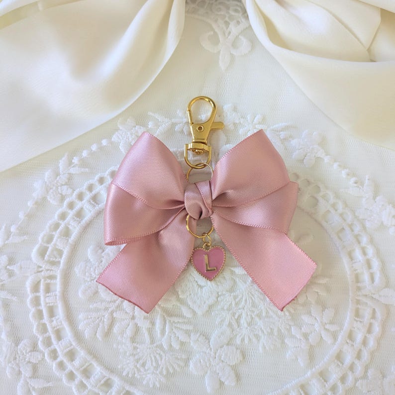 Peut inclure: Un porte-cl&eacute;s en forme de n&oelig;ud rose en satin, avec un fermoir dor&eacute; et un charm en forme de c&oelig;ur. Le charm porte la lettre "L". Le n&oelig;ud est nou&eacute; de mani&egrave;re classique.