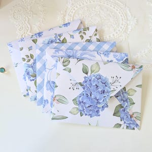 May include: A collection of patterned envelopes with floral designs. The envelopes display a range of patterns, including blue hydrangea, roses, and gingham, set against a white backdrop. The colour scheme is predominantly blue and white, producing a delicate, refined appearance.