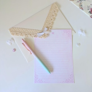May include: A pink and white lined stationery sheet with a decorative border and a light blue and pink pen on top of a white envelope with lace trim.