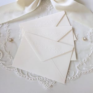 May include: A stack of three cream-colored envelopes featuring a delicate embossed vine and heart pattern along the triangular flap. The envelopes are made of heavy cardstock and have a smooth, matte finish, presenting a clean and uniform appearance.