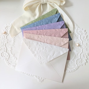 Könnte beinhalten: Eine Sammlung pastellfarbener Umschläge mit geprägten Blumen- und Schmetterlingsmustern. Die Umschläge sind fächerförmig angeordnet und zeigen eine Vielzahl von Farben, darunter Weiß, Rosa, Lila und Blau. Eine weiße Schleife und eine Spitzentrimmung befinden sich im Hintergrund.