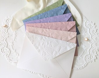 May include: A collection of pastel-colored envelopes with embossed floral and butterfly designs. The envelopes are arranged in a fan shape, showcasing a variety of colors including white, pink, purple, and blue. A white bow and lace trim are in the background.
