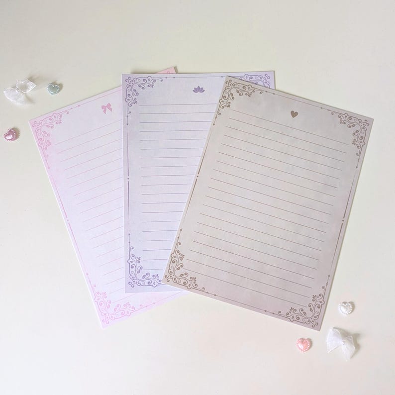 May include: Three sheets of lined stationery paper with decorative borders. The paper is in shades of pink, purple, and beige.