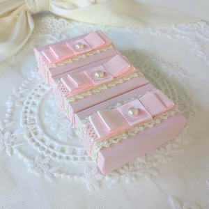 May include: Three rectangular light pink boxes arranged in a row. Each box is decorated with a strip of cream-colored lace trim, a light pink satin ribbon bow, and a single white faux pearl bead centered on the bow.