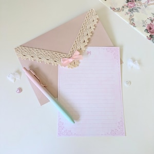 May include: A pink and white lined stationery set with a delicate lace trim and a pink bow. The set includes a pink envelope and a pen with a pink and green ombre finish.