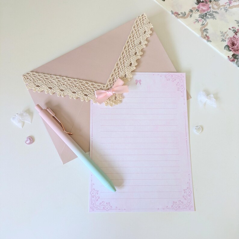 May include: A pink and white lined stationery set with a delicate lace trim and a pink bow. The set includes a pink envelope and a pen with a pink and green ombre finish.