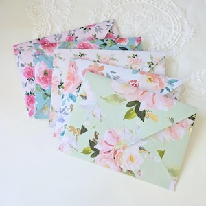 May include: A collection of floral-patterned envelopes in various colors. The envelopes feature watercolor designs of pink and peach roses, with green and blue foliage. The envelopes are arranged in a fan shape, showcasing the different patterns.
