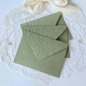 May include: Sage green envelopes with embossed floral patterns. The envelopes are arranged on a white lace doily, with a cream-colored ribbon and a decorative jewel in the background. The envelopes are a muted green color.