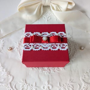 Könnte beinhalten: Eine rote Geschenkbox, verziert mit weißer Spitzenborte, rotem Band und einer perlenbesetzten Verzierung. Die Box steht auf einer weißen Spitzentischdecke, im Hintergrund ein cremefarbener Bogen. Zwei kleine Perlenakzente sind sichtbar.
