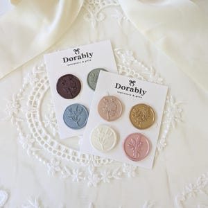 May include: Two cards of Dorably stationery and gifts wax seals. Each card has three circular seals with floral designs in various colors, including brown, blue, gold, pink, and white. The cards are on a white lace surface.
