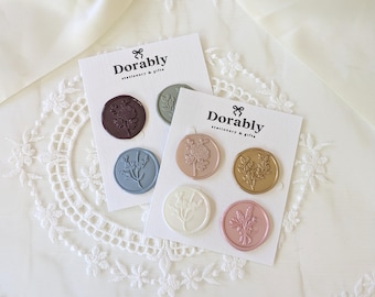 May include: Two cards of Dorably stationery and gifts wax seals. Each card has three circular seals with floral designs in various colors, including brown, blue, gold, pink, and white. The cards are on a white lace surface.