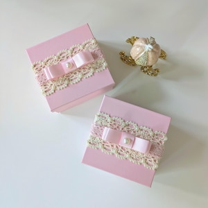 May include: Two pink gift boxes with ivory lace trim and pink satin bows, each adorned with a pearl. A decorative, gold-toned pumpkin carriage with pink and crystal accents sits nearby. The boxes are ideal for small gifts.