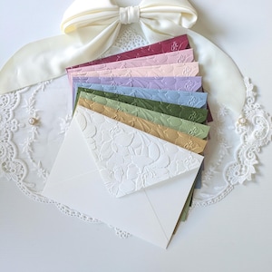 Puede incluir: Una pila de sobres multicolores con un patrón floral en relieve en la solapa triangular. Los colores incluyen blanco, dorado, verde oliva, azul claro, lavanda, rosa claro y magenta intenso, dispuestos en forma de abanico.